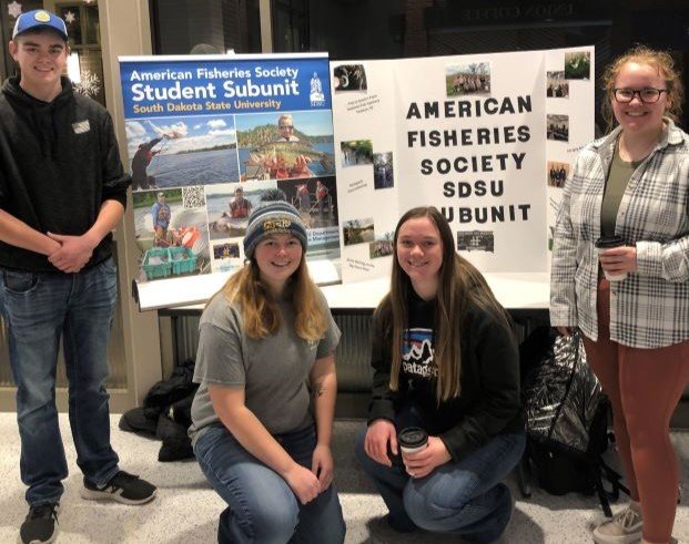 American Fisheries Society Student Subunit pose in front of signs that contain information about their club at the hot chocolate social.