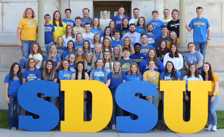 Group of admissions ambassadors all wearing yellow and blue t-shirts standing in front of the campanile with a SDSU sign.