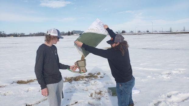 Pheasants Forever Club spread a restoration seed mix over freshly fallen snow to help the seeds chances of success. one student pours seed into a handheld seed spreader for another student.