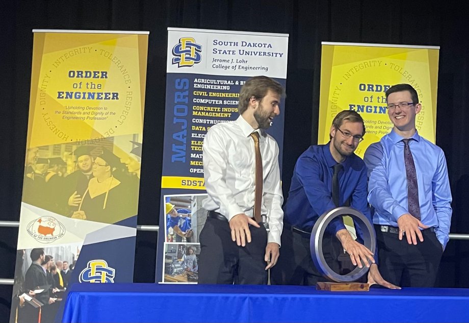 Class of 2023 graduates have a little fun showing off their Order of the Engineer ring at the May 5 ceremony. Pictured, from left, are Christian Fuller, Cody Decker and Tyler Fogelson. Decker puts his hand through the tabletop model that was created this year by Tyler Hanks, head of the production lab in Chicoine Architecture, Mathematics and Engineering Hall.
