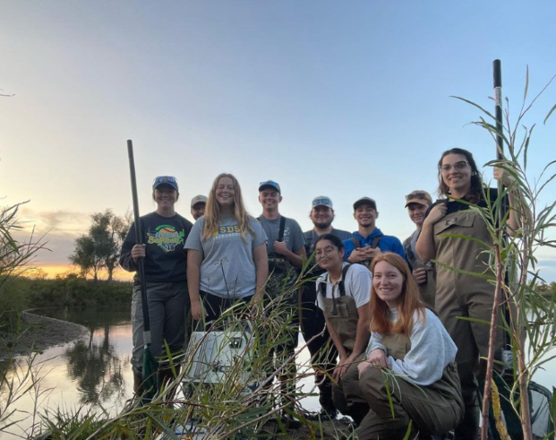 Ten members of the American Fisheries Society Student Subunit at the lake electrofishing while wearing waders and holding a net.
