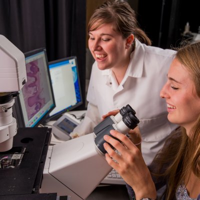 Two women work in a lab, one with a microscope
