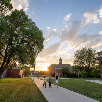 Shot of two students walking on campus with blue skies and the sun coming up over the buildings.