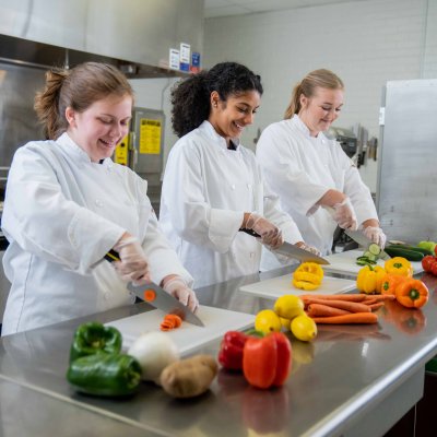 three female students in white lab coats cutting vegetables
