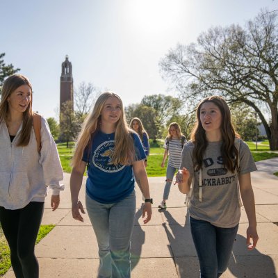 Three students walking on campus with the campanile in the background.