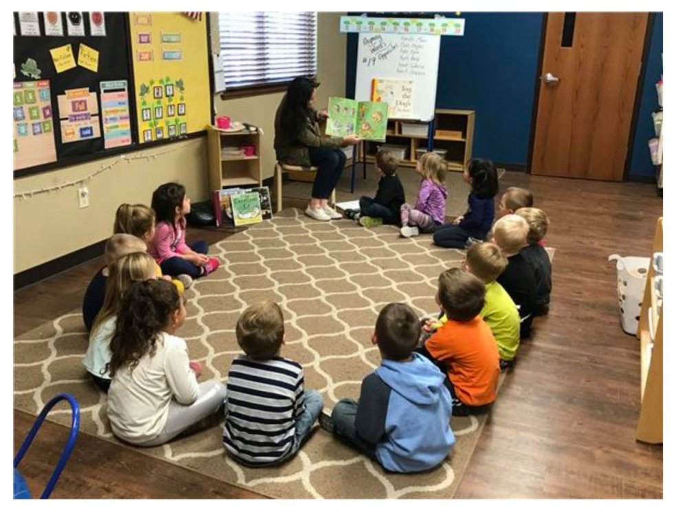 Teacher reads book to group of young students sitting in a circle on the floor.