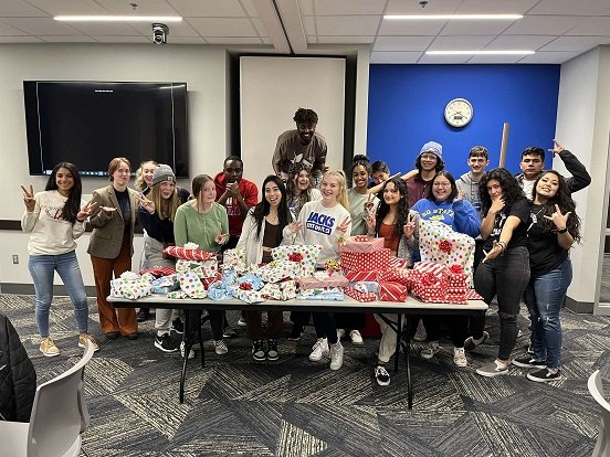 Students standing in a big group making silly faces in front of a table full of wrapped gifts 