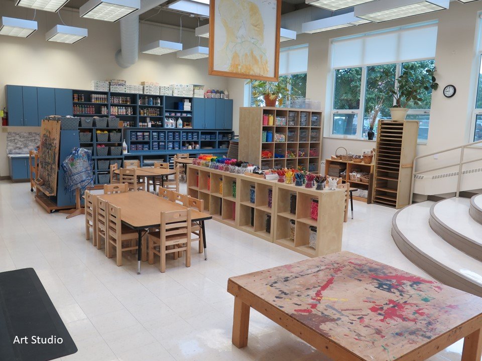 The art studio that includes multiple tables and chairs as well as shelving units full of materials to engage with and create.