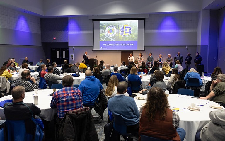 An event with several people seated at tables and a presentation saying "Welcome SFSD Educators" on the screen.
