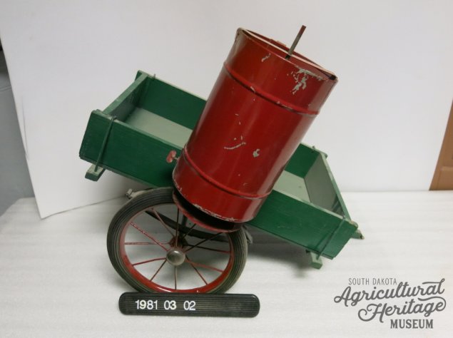 SDAHM Collection, 1981:003:002.  Red cylindrical bin attached to a green square structure.  The green wooden base is attached to two wheels.