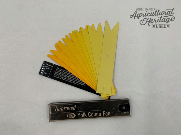 2023:022:01 Egg yolk color fan that pivots in the corner.  The cover is black with the words "Improved Roche Yolk Color Fan".  There are nine other cards in the fan in various shades of yellow with a number printed on each card.  The top card is the palest and has the number "1" printed on it.  The fan fits in a clear plastic case that has yellowed with age.
