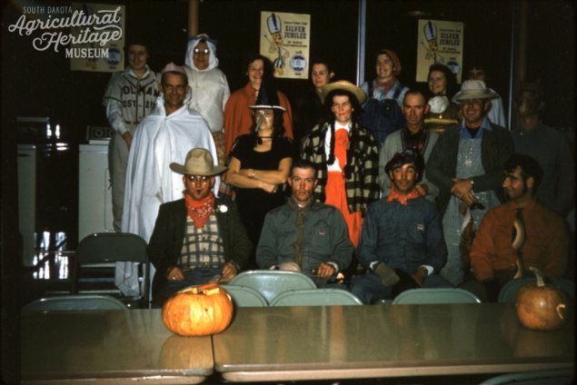 2003.027:0087.  Color photo of 17 people dressed in costume at a Halloween party.  Some of the identifiable costumes include a witch, farmer, and baseball player.