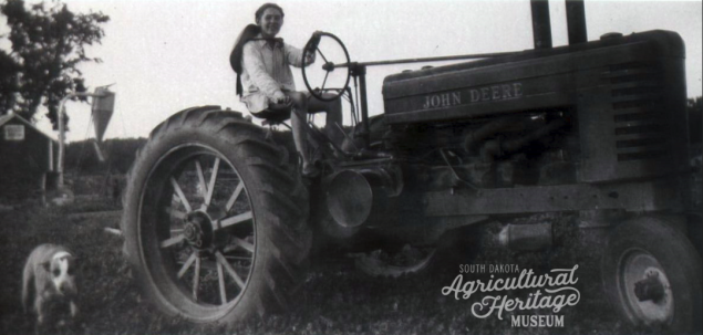 Black and white photo of a woman riding a John Deere tractor.  To the left of the tractor is a dog.