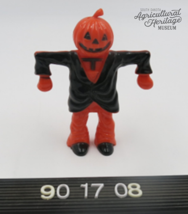 Donated by Roger Knapp, 1990:017.  This image shows an orange scarecrow with a carved jack-o-lantern head wearing a black coat and black shoes.  Below the scarecrow are the numbers "90 17 08".