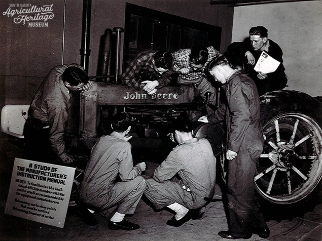 A group of young men are standing and crouched around a tractor preforming maintenance on it 