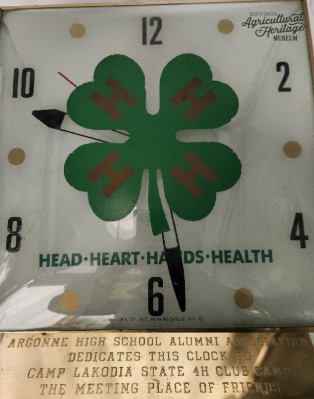 2001:118:002.  Clock with white background, even numbers printed in black, gold dots for odd numbers, and a 4-H clover logo in the middle.  Under the clover reads "HEAD - HEART - HANDS - HEALTH".  Below the clock hangs a gold plaque with that reads “Argonne High School Alumni Association dedicates this clock to Camp Lakodia State 4H Camp the meeting place of friends.”