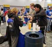 Three chemistry club students talking to two students and performing an experiment while standing at table during a middle school outreach fair.