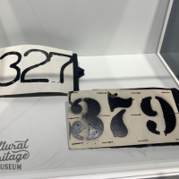 SDAHM 2021:032:017 & 2021:032:018.  Two arm bands that are made of white cardboard.  The band on the upper left side has the number 327 printed in black ink, while the band on the lower right side has the number 379 in black ink.