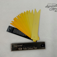 2023:022:01 Egg yolk color fan that pivots in the corner.  The cover is black with the words "Improved Roche Yolk Color Fan".  There are nine other cards in the fan in various shades of yellow with a number printed on each card.  The top card is the palest and has the number "1" printed on it.  The fan fits in a clear plastic case that has yellowed with age.