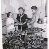 Leland Sudlow Photograph Collection.  A black and white image of three women in a kitchen judging 37 cherry pies.  Some of the pies are on two metal carts while one of them is being held by the women in the center of the photograph.