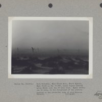 Black and white photo of a man walking through a dust storm.  There are drifts of soil surrounding the man on the ground.  The photo is backed on a gray paper that reads "Serial No. 33-202:- Iowa Township, Shue Creek Area, South Dakota. Same field as No. 33-201, human figure (Caird) going South into the 26 mile wind.  Small drifts can be seen, in the foreground of the picture, forming on the protected side of each Russian Thistle. (10/15/35)"