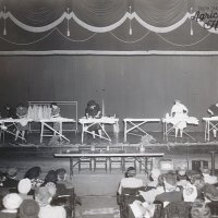 Leland Sudlow Collection 70-9-2.  A black and white photo of five women up on a raised stage ironing shirts.  There are multiple people seated in the audience.