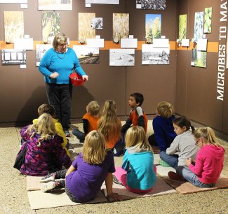 A group of young children on sitting on mats at the Agricultural Heritage Museum while the presenter is speaking at the microbes exhibit.